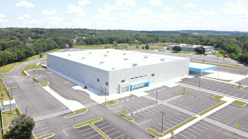 Aerial view of a large, white industrial warehouse with a flat roof, surrounded by an expansive, mostly empty parking lot. The building is set against a backdrop of green trees and open fields under a partly cloudy sky.
