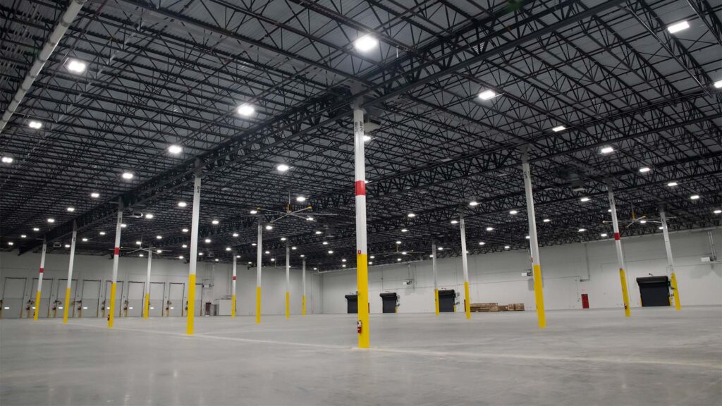 Interior of a large, empty warehouse with high ceilings and yellow pillars. The floor is concrete, and the space is well-lit with numerous overhead lights. Several doors and dock areas line the walls.