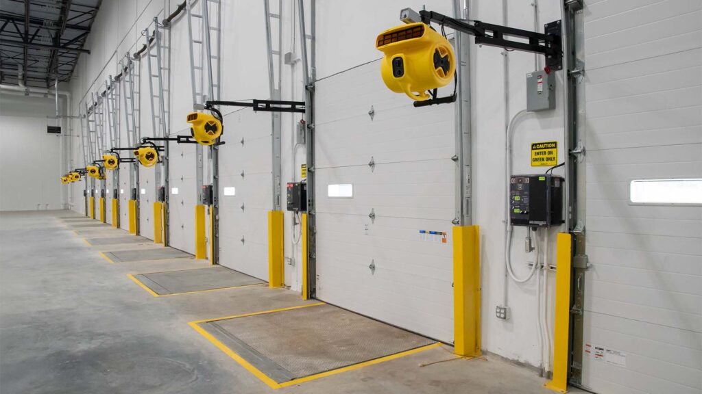 Interior of a warehouse loading dock with multiple closed white garage-style doors. Each door has a yellow loading dock light and bumper guards. The area is clean and well-lit, with concrete floors and high ceilings.