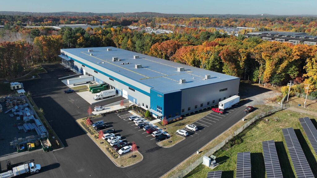 Aerial view of a large blue and white industrial building surrounded by parking lots with cars and trucks. The building is bordered by autumn-colored trees, and solar panels are visible in the foreground.
