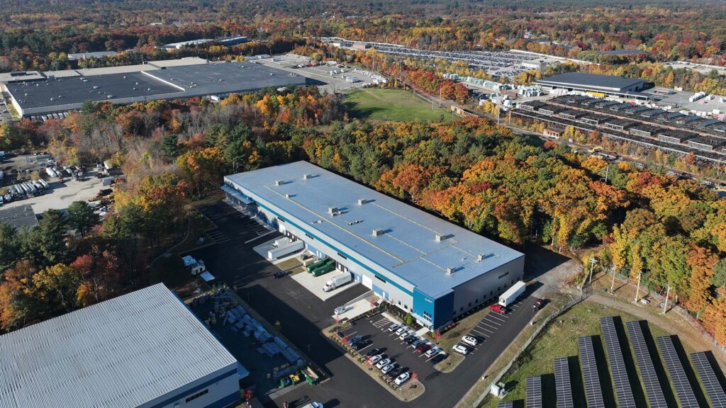 Aerial view of industrial buildings surrounded by autumn trees with colorful leaves. Several large warehouses and parking areas are visible, alongside open plots of green land. Solar panels line the foreground, and a network of roads connects the facilities.