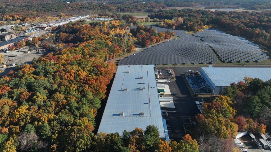 Aerial view of a large industrial facility with solar panels on the roof and adjacent ground. The surrounding landscape features dense autumnal forests with trees in shades of orange, yellow, and green. Buildings and roads are visible in the distance.