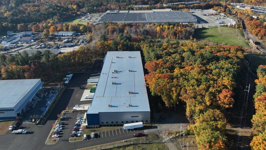 Aerial view of a large industrial complex surrounded by autumn-colored trees. Several rectangular warehouse buildings with parking lots are visible. Roads and a few smaller structures are also present in the landscape.