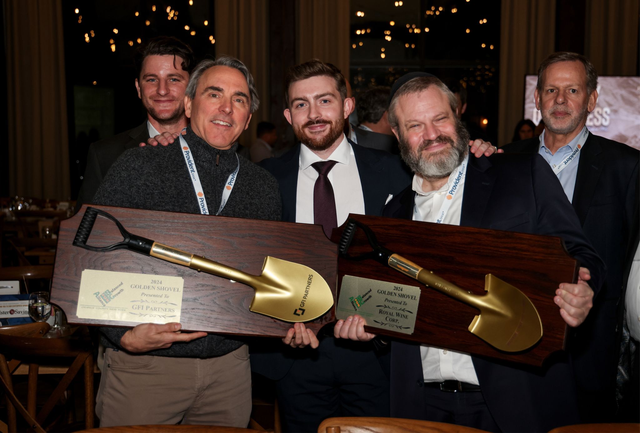 Five men stand together indoors, smiling. Two of them hold large wooden plaques with gold shovels mounted on them. The room is warmly lit, and people are visible in the background, suggesting a festive or celebratory event.