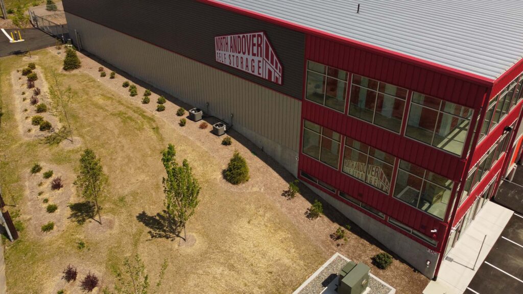 A large red and gray industrial building with a sign that reads Storage City on the side. The building has multiple windows, and theres a paved parking area next to it. The surrounding area is landscaped with grass and small trees.