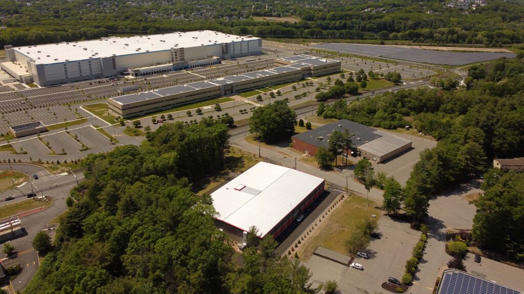 Aerial view of a large industrial complex with several rectangular buildings surrounded by parking lots and greenery. Solar panels are visible on one of the building roofs and more are installed on the ground nearby. Trees and a road border the area.