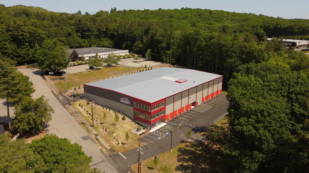 Aerial view of a large warehouse building surrounded by trees and greenery. The building has a gray roof, red trim, and a parking lot with a few cars. There are other industrial buildings and a forested area in the background.