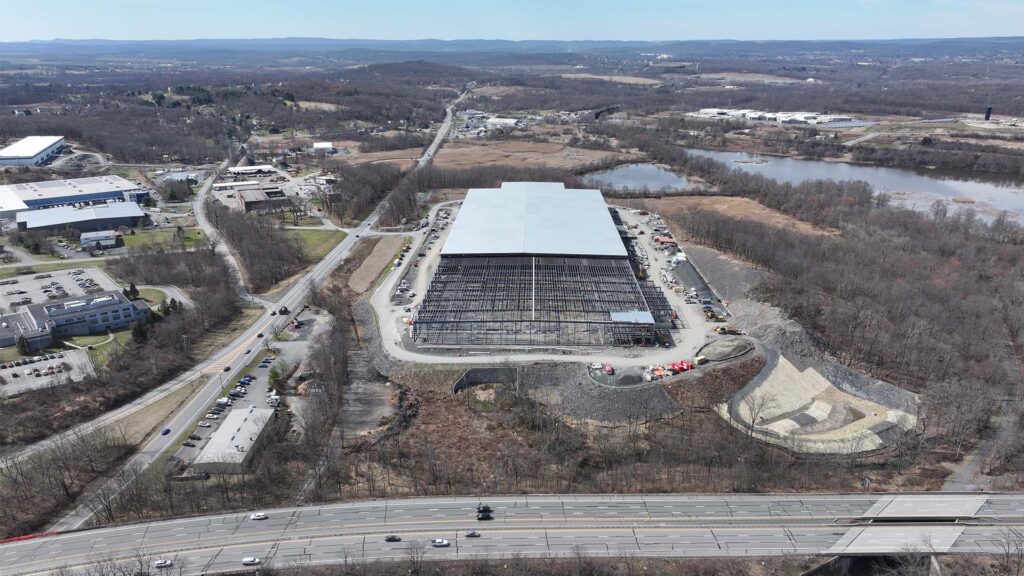 Aerial view of a construction site with a large industrial building being developed, surrounded by roads and scattered buildings. The structure is partially completed with a visible steel framework, set amidst a wooded landscape near a river.