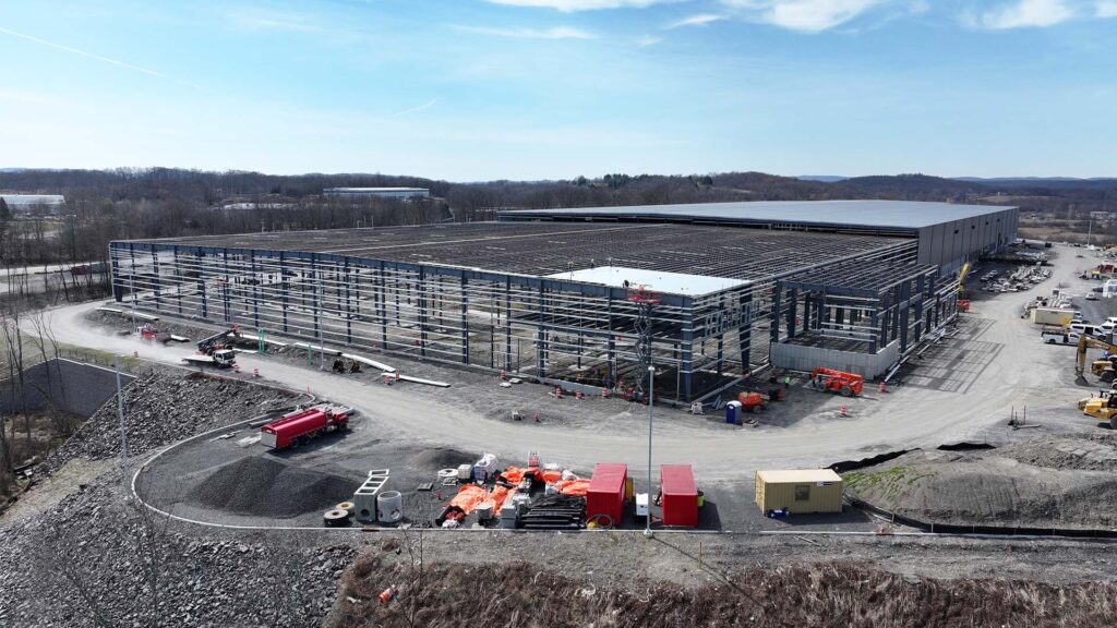 A large industrial building is under construction, with a steel frame partially completed. Construction vehicles and materials, including red containers and piles of gravel, are scattered around the site. The background shows a wooded area and a clear sky.