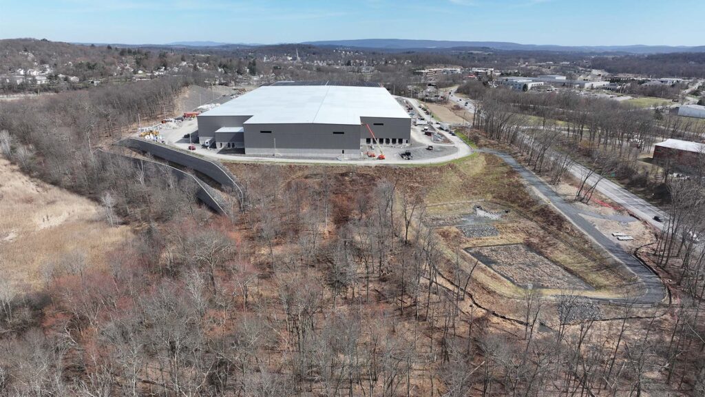 A large industrial building surrounded by trees with a winding road leading to it. The landscape includes cleared areas, with construction vehicles visible near the building. In the distance, hills and a small town are visible under a clear blue sky.