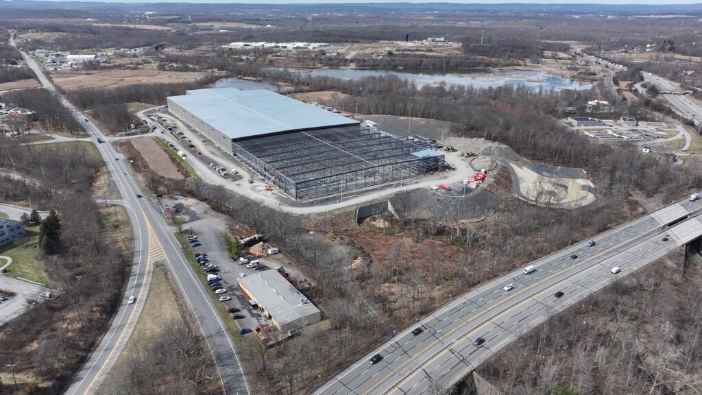 Aerial view of a large construction site featuring a partially built structure with a blue roof and visible steel framework. Surrounding the site are roads, wooded areas, and nearby highways under clear skies.
