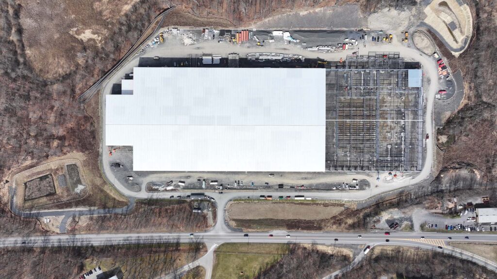 Aerial view of a large industrial building with a white roof, partially under construction. Surrounding areas include trees, construction equipment, and a road with vehicles at the bottom of the image.