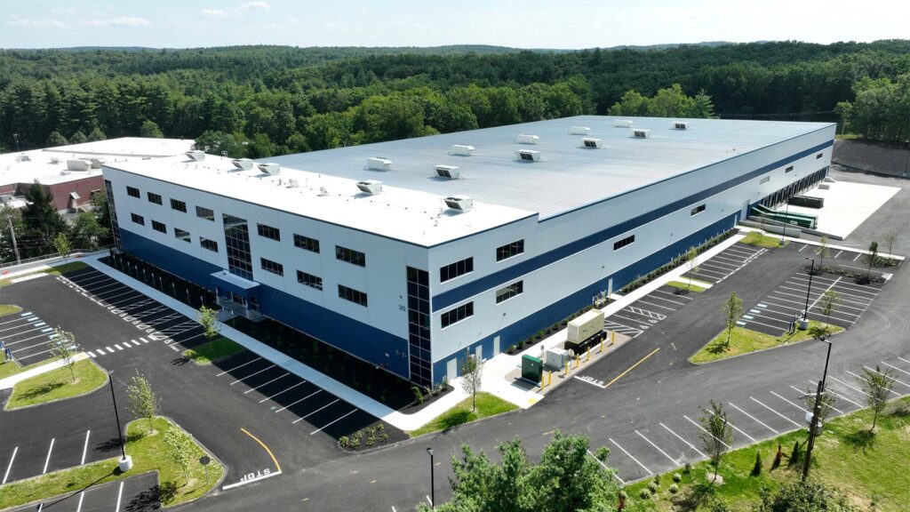 A large blue and white warehouse building surrounded by empty parking spaces. The area is lush with green trees and clear skies, suggesting a sunny day. The building has multiple loading dock doors and windows.