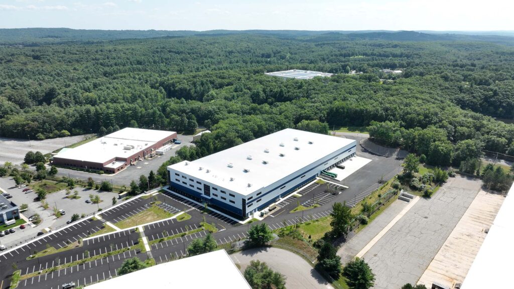 Aerial view of a large industrial complex surrounded by dense forest. The complex features two main buildings with white roofs and ample parking spaces. The surrounding landscape is lush and green under a clear sky.