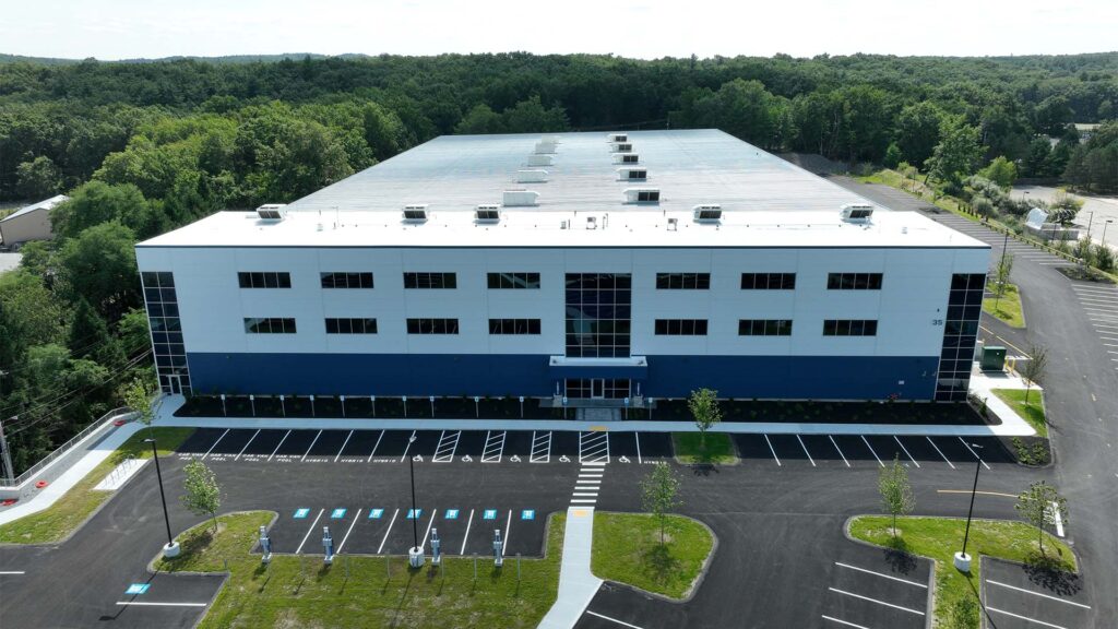 Aerial view of a large, rectangular building with a flat roof, surrounded by a parking lot and green foliage. The building has a blue and white exterior, with a main entrance at the front and trees lining the surroundings.