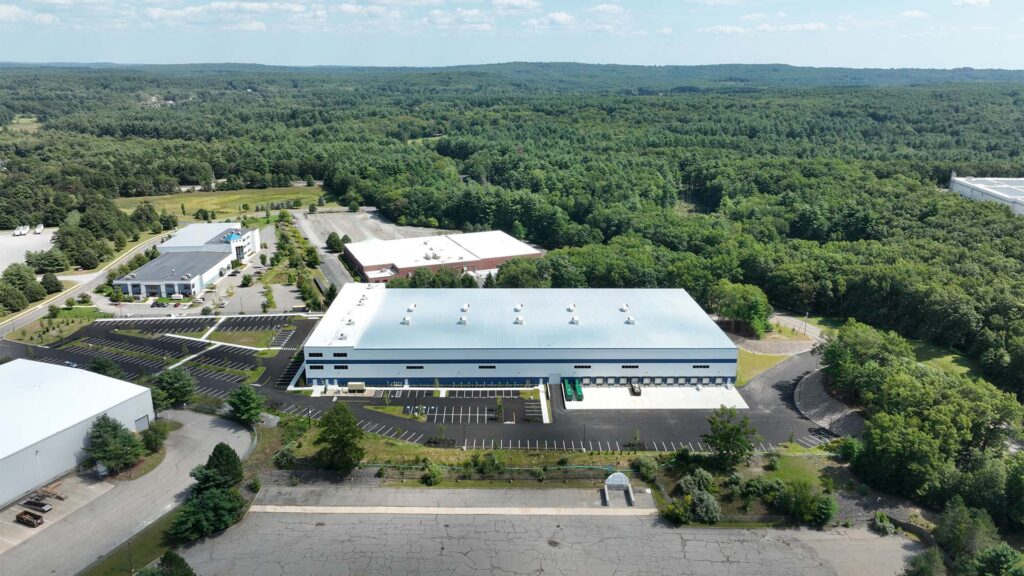 Aerial view of a large industrial building with a white roof surrounded by trees and parking lots. Several smaller buildings are nearby. The area is green and wooded, indicating a rural or suburban location.