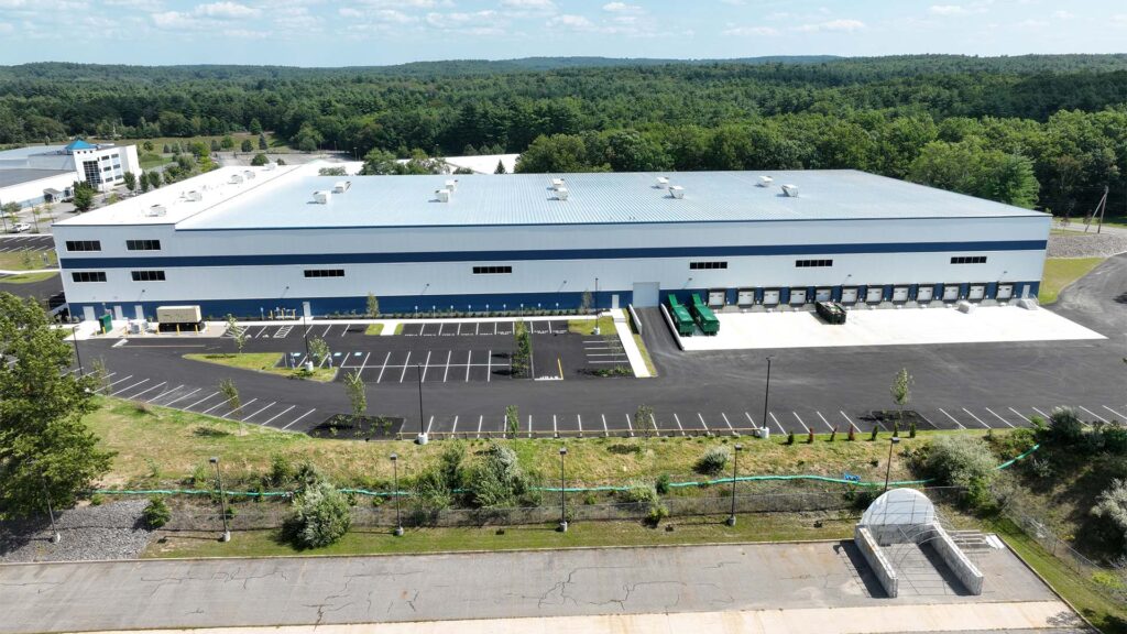Aerial view of a blue and white warehouse surrounded by a parking lot, loading docks, and landscaped greenery. Forested area in the background under a clear blue sky.
