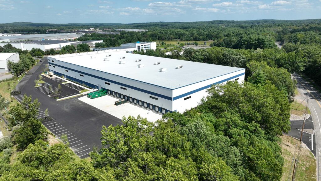 Aerial view of a large rectangular warehouse surrounded by greenery. The building has a white exterior with blue accents and multiple loading dock doors. A parking lot with lined spaces is visible along one side. Hills and trees extend in the background.