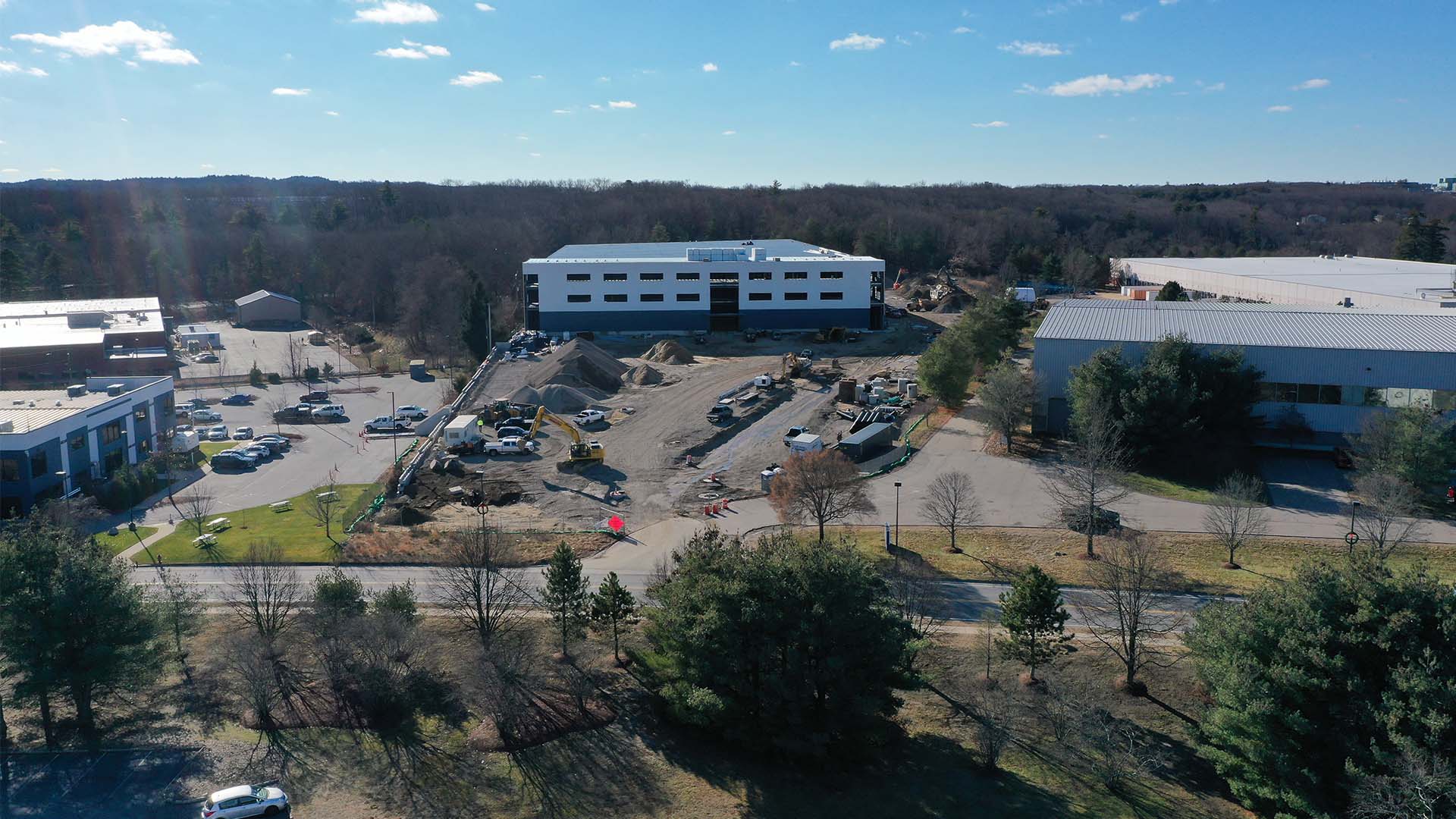 Aerial view of a construction site with a partially completed building surrounded by trees and parked vehicles. Other industrial buildings are visible nearby under a clear blue sky.