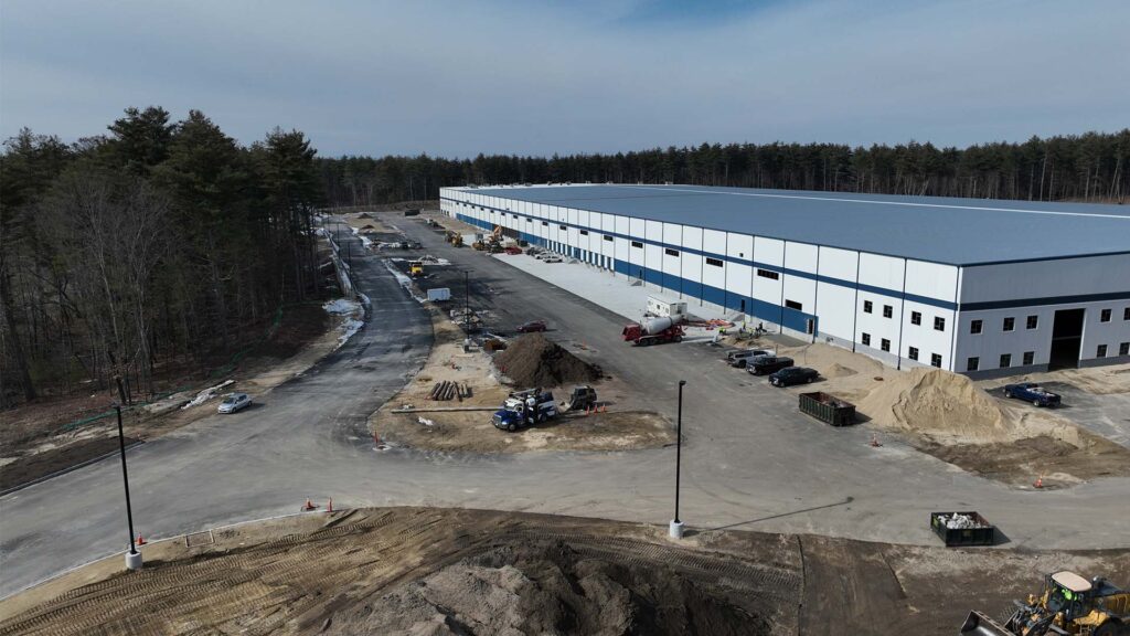 Aerial view of a large warehouse with a blue and white exterior surrounded by forest. The site has a parking area with several vehicles and ongoing construction work with soil piles and equipment scattered around.