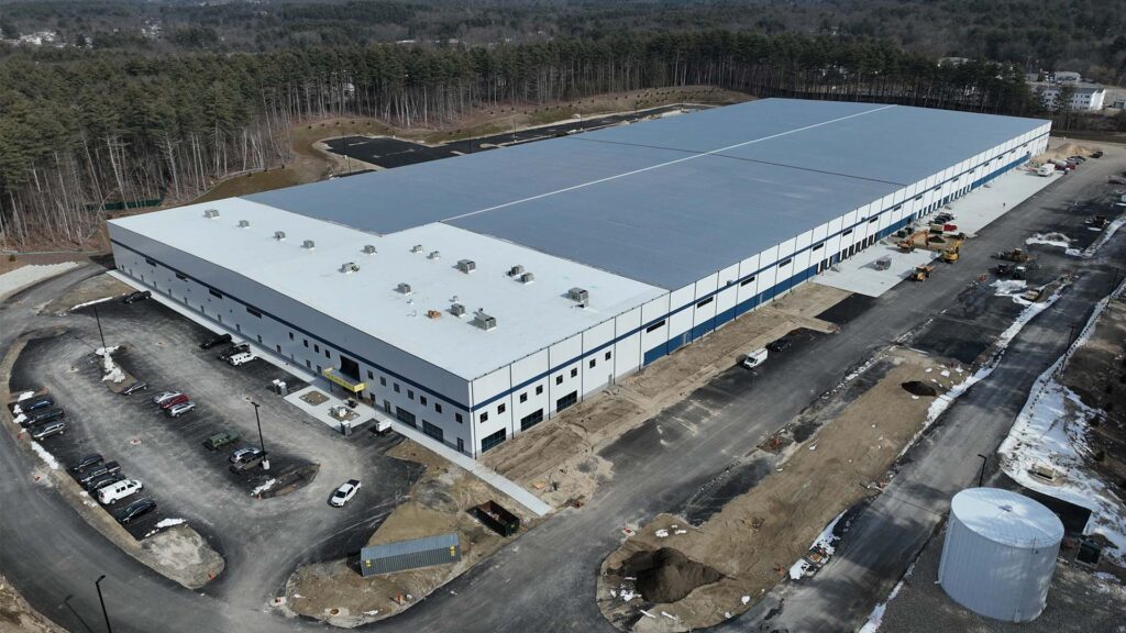 Aerial view of a large warehouse with a flat roof, surrounded by a partially completed parking lot and construction vehicles. The building is set against a backdrop of trees and a forested area. Snow patches are visible on the ground.