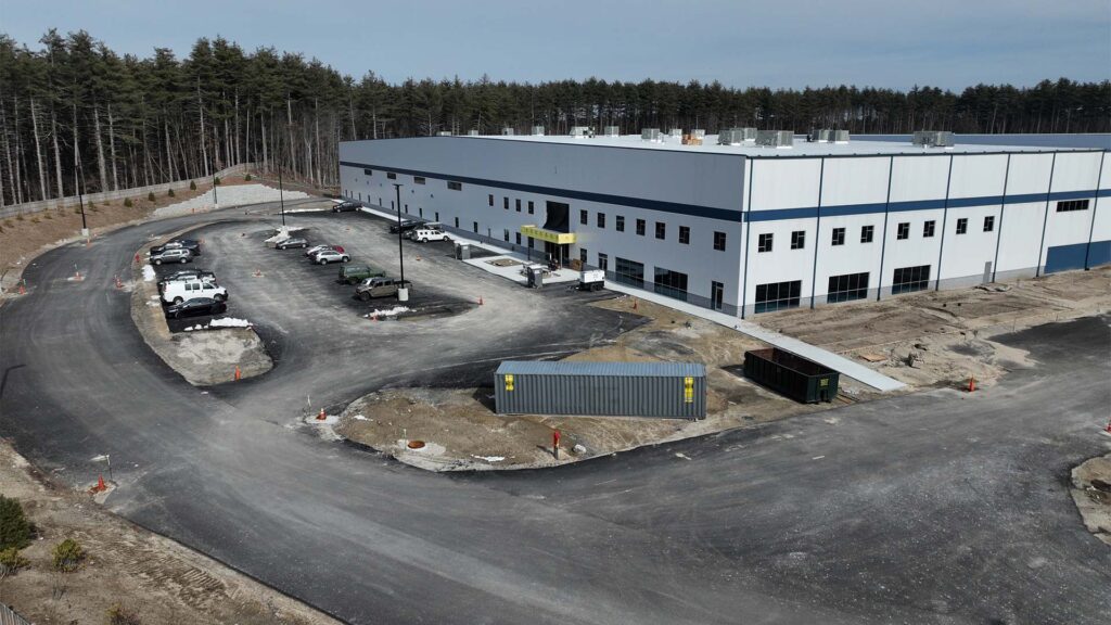 Aerial view of a large, rectangular white industrial building with blue accents. Surrounding it is a paved road loop and several parked vehicles. The scene is bordered by a wooded area under a cloudy sky. A shipping container is visible near the building.
