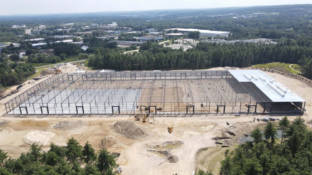 Aerial view of a large construction site with a partially built steel frame structure surrounded by forested areas and industrial buildings in the background. Construction equipment and dirt mounds are visible on the ground.