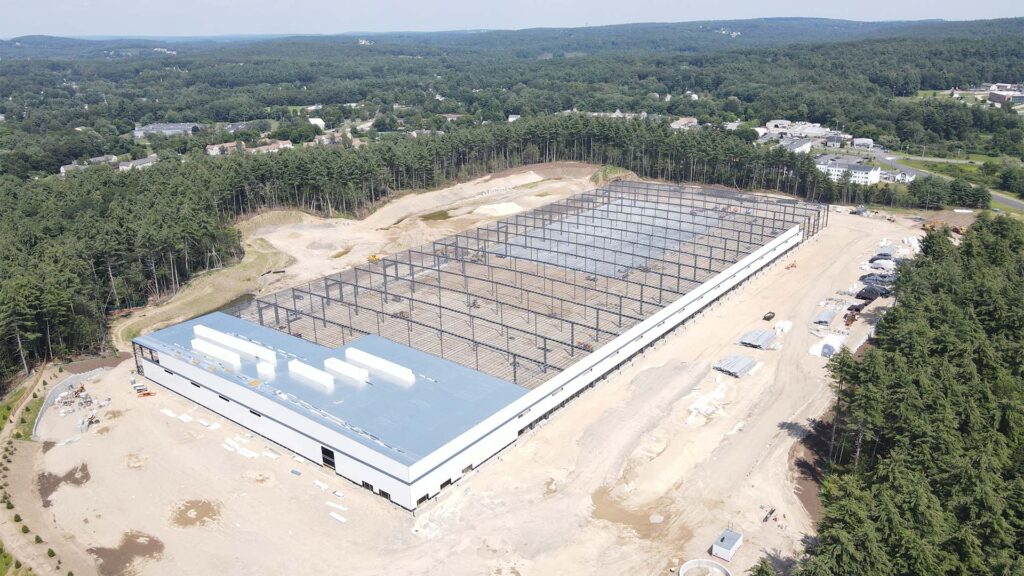 Aerial view of a large industrial building under construction, surrounded by trees. The rectangular structure has a metal frame and partial roofing. Construction materials and machinery are visible around the site, set within a forested area.