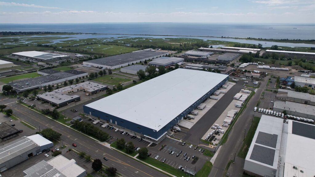 Aerial view of an industrial park near a body of water. Large warehouses with parking lots and trucks are visible, surrounded by greenery and roads. A coastline with water is in the distance under a partly cloudy sky.