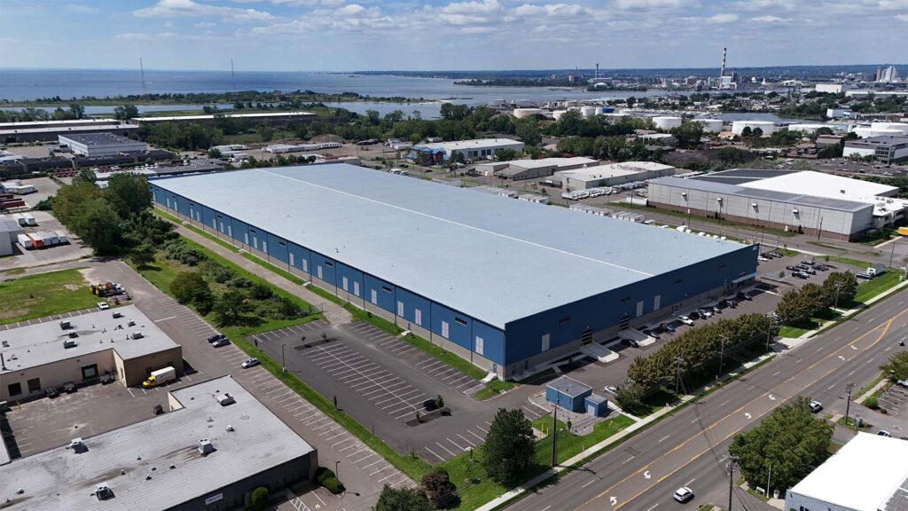 Aerial view of a large blue warehouse surrounded by parking lots and green spaces. Nearby are other industrial buildings, roads, and a body of water in the background under a partly cloudy sky.