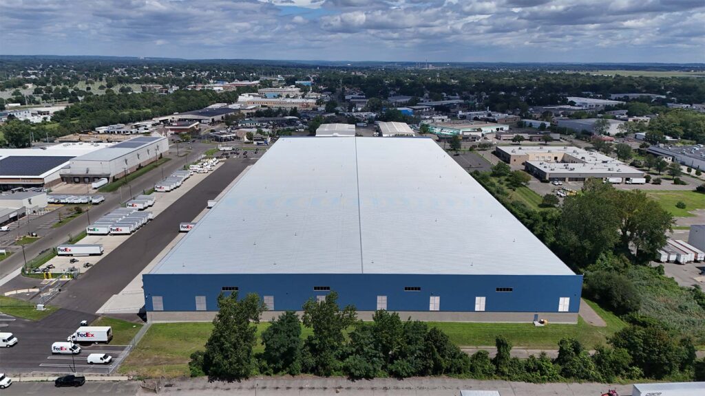 Aerial view of a large blue and white warehouse in an industrial area. Surrounding buildings, roads, and greenery are visible under a partly cloudy sky. Trucks and cars are parked near the warehouse.