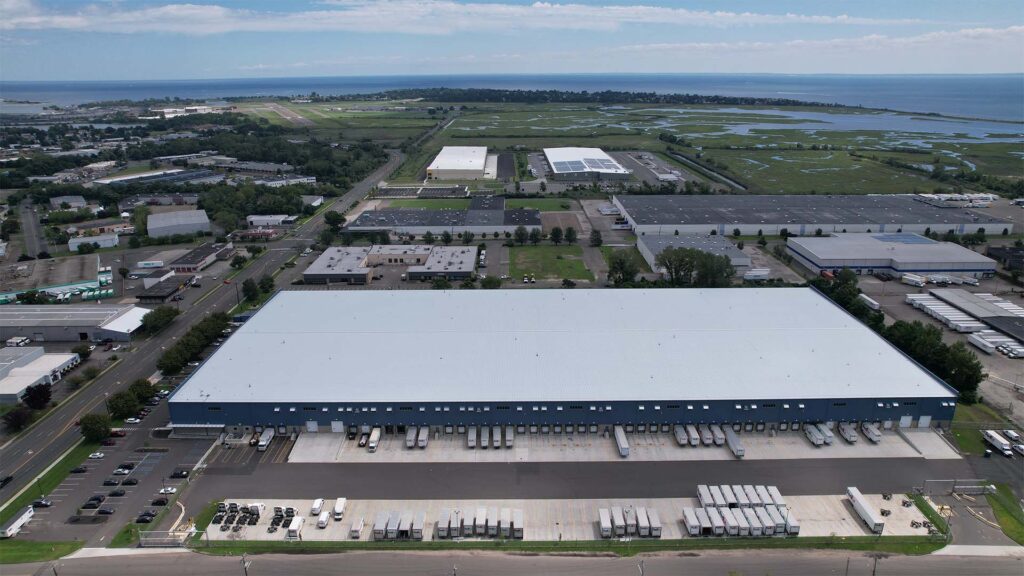 Aerial view of a large industrial building with a light blue roof and multiple loading docks. Surrounding the building are parking lots with numerous trucks. The area is an industrial park with other warehouses, roads, and distant greenery along the coast.