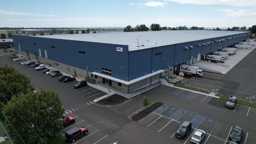 A large industrial warehouse with a blue exterior and FedEx logos on loading docks. Several FedEx trucks are parked at docking stations. The building is surrounded by a parking lot with cars and trees. The sky is mostly clear with some clouds.