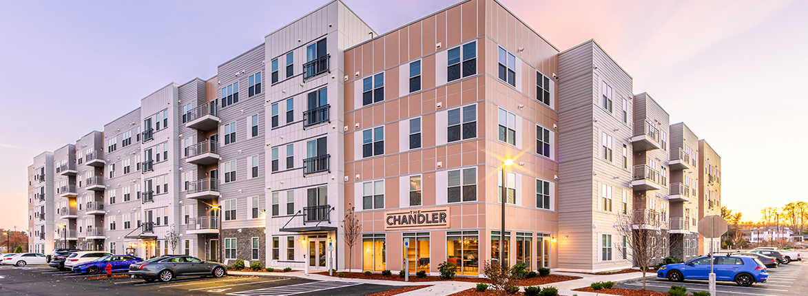 A modern, multi-story apartment building with a mix of beige and gray siding. The entrance has the sign CHANDLER, and the building is surrounded by a parking area with a soft evening sky in the background.