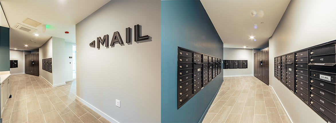 A modern mailroom with beige tiled floors and blue-gray walls. Multiple rows of black mailboxes line the walls. A sign with the word MAIL is mounted on one of the walls. Bright, even lighting illuminates the space.