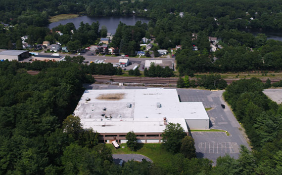 Aerial view of a large, flat-roofed industrial building surrounded by trees. A railway line runs parallel to the building. Nearby, theres a small neighborhood with houses and a distant body of water. Parking lot in the foreground is mostly empty.