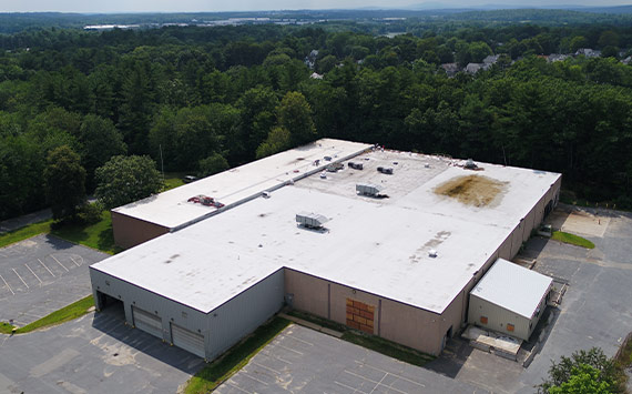Aerial view of a large warehouse building with a white flat roof, surrounded by a parking lot. The building is set amidst a forested area with a distant view of hills and a clear sky.