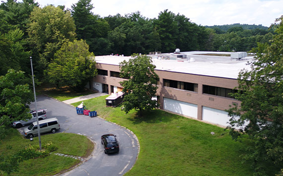 Aerial view of a rectangular, two-story brick building with a flat roof, surrounded by trees. Two vehicles are on a curved driveway leading to the building. Theres a trash bin area nearby, and the scene is set under a partly cloudy sky.