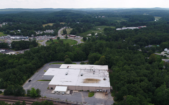 Aerial view of a large, white industrial building surrounded by dense green forest. In the background, a road curves through a residential area with houses. The sky is cloudy, and the landscape is mostly verdant with patches of development.