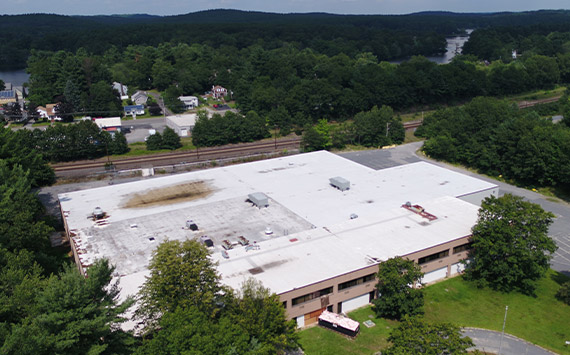 Aerial view of a large, flat-roofed industrial building surrounded by trees. The roof shows wear in spots. Nearby are train tracks, small structures, and a river, set against a backdrop of dense forest and distant hills.