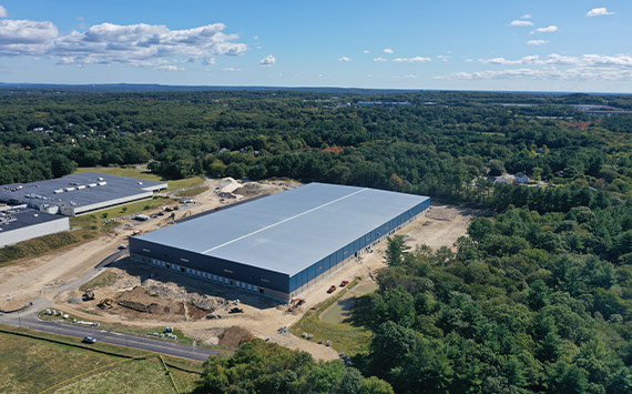 Aerial view of a large rectangular warehouse under construction, surrounded by greenery and trees. Nearby, there are roads and smaller buildings. The sky is clear with a few clouds, indicating a sunny day.