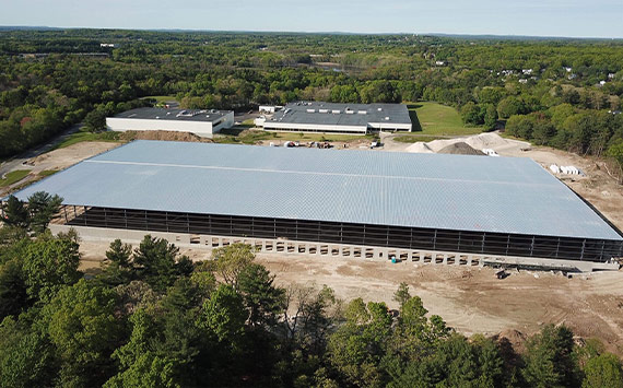 Aerial view of a large rectangular warehouse under construction, surrounded by a mix of cleared land and dense green trees. Nearby, there are several smaller completed buildings, and the landscape extends into a distant horizon of more trees.