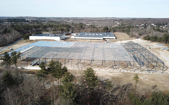 Aerial view of a large construction site with a steel framework partially erected. Surrounding the site are trees and a few buildings in the background under a clear sky.