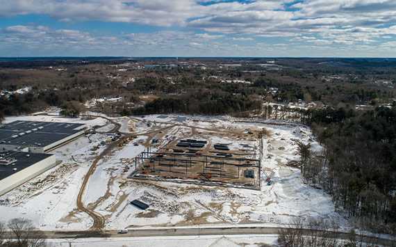 Aerial view of a snowy landscape showing a construction site with steel beams and equipment. Surrounding the site are patches of snow-covered terrain, a few roads, and a nearby industrial building. The sky is cloudy, with scattered patches of blue.