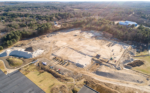 Aerial view of a construction site surrounded by trees. The site features several large piles of dirt and sand, with construction vehicles scattered throughout. A few buildings are visible in the distance, and the area is bordered by a forested landscape.