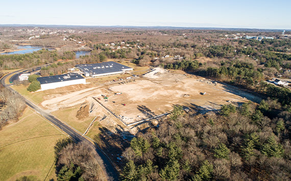 Aerial view of a large construction site next to an industrial building, surrounded by trees. The area is mostly cleared, with some equipment visible. In the background, a river and distant town are visible under a clear sky.