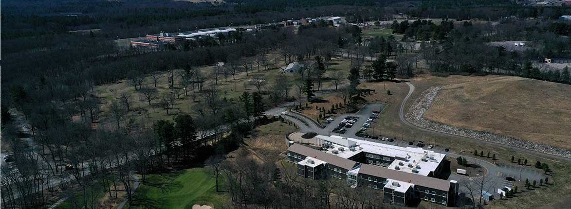 Aerial view of a complex with multiple dark-roofed buildings surrounded by trees and grassy areas. Parking lots and roads are visible, with additional buildings in the background, set in a wooded landscape.