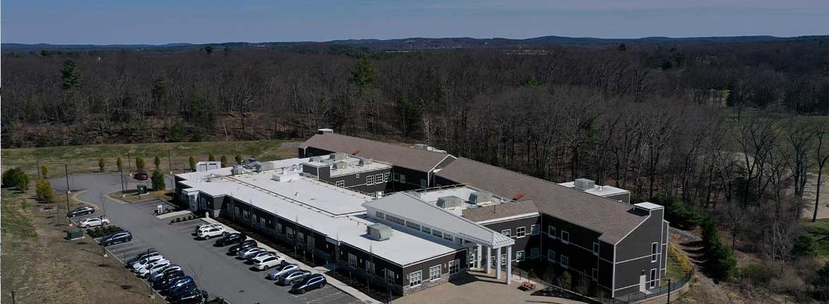 Aerial view of a large, single-story building with a white roof surrounded by a parking lot with parked cars. The building is set against a backdrop of trees and rolling hills under a clear sky.