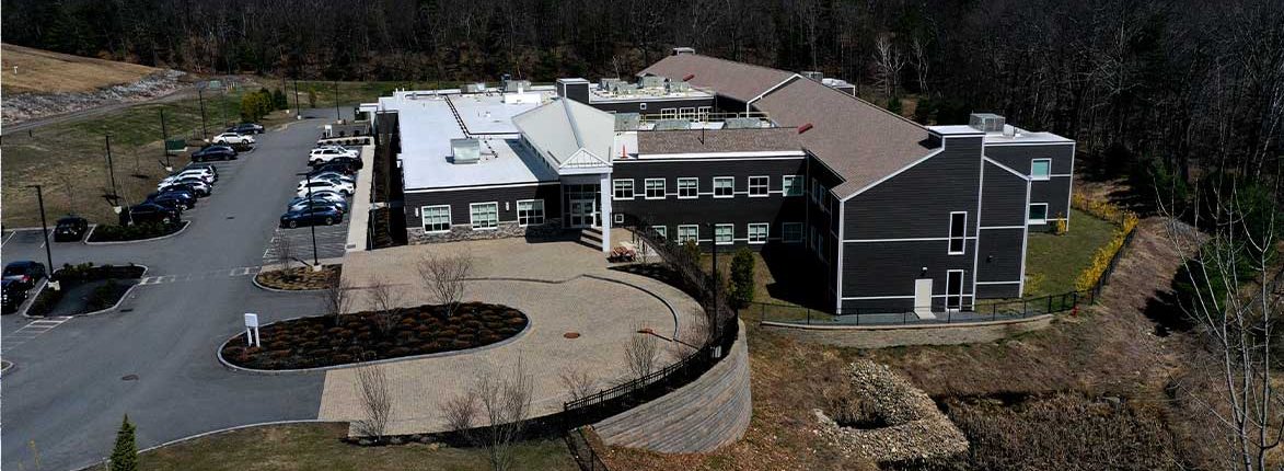Aerial view of a large, modern building surrounded by trees and a parking lot with several cars. The building has a mix of white and dark siding, flat roofs, and a curved entrance area with landscaped shrubs. Parking spaces are visible to the left.