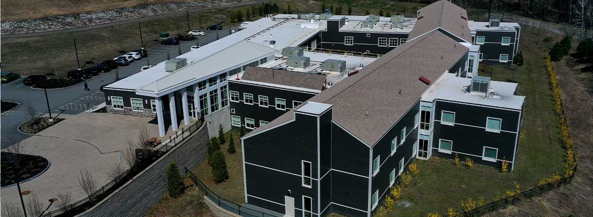 Aerial view of a dark green educational building with a tan roof, featuring white trim and multiple windows. The structure is surrounded by landscaped paths, parking areas, and trees.